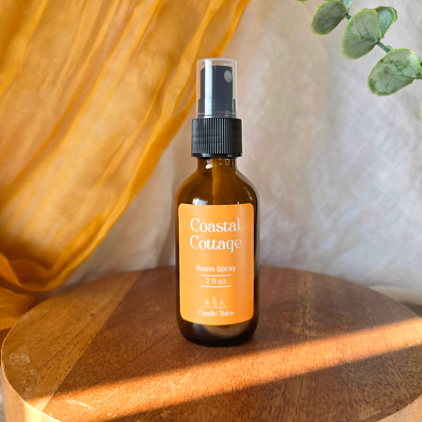Bottle of Coastal Cottage room spray on a wooden surface with an orange drape and faux plant in the background.