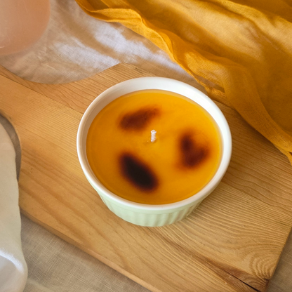 A creme brulee candle in a green ceramic container on a wooden cutting board with an orange cloth in the background.