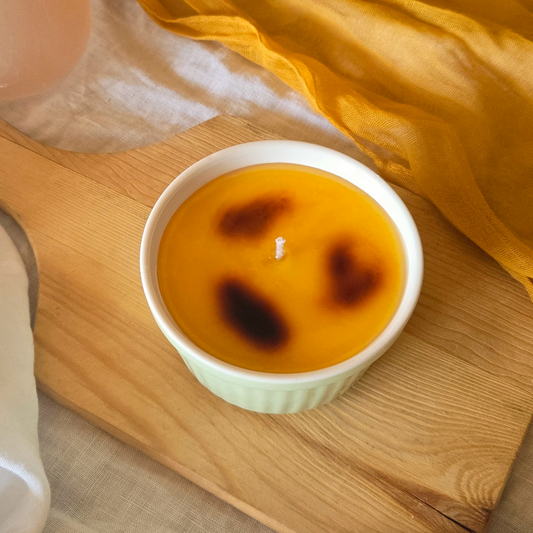 A creme brulee candle in a green ceramic container on a wooden cutting board with an orange cloth in the background.