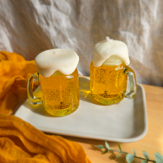 Two beer candles in mini mason jars with foam on top, sitting in a grey tray, surrounded by yellow fabric and green leaves.