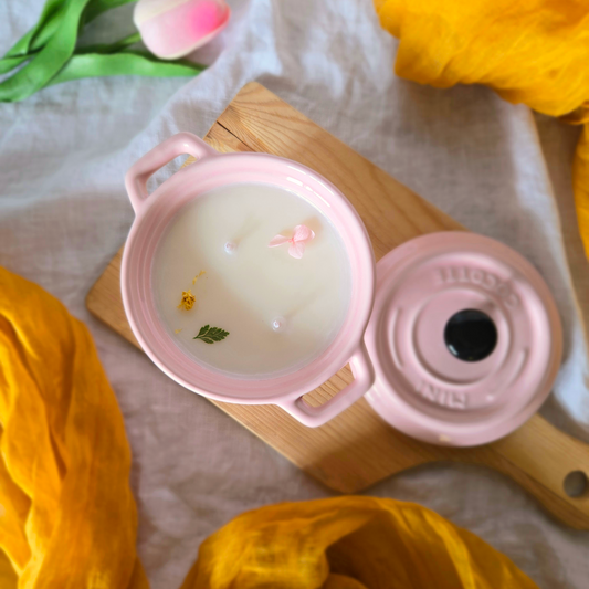 Pink ceramic pot soy wax candle on a wooden board, surrounded by orange fabric and flowers.